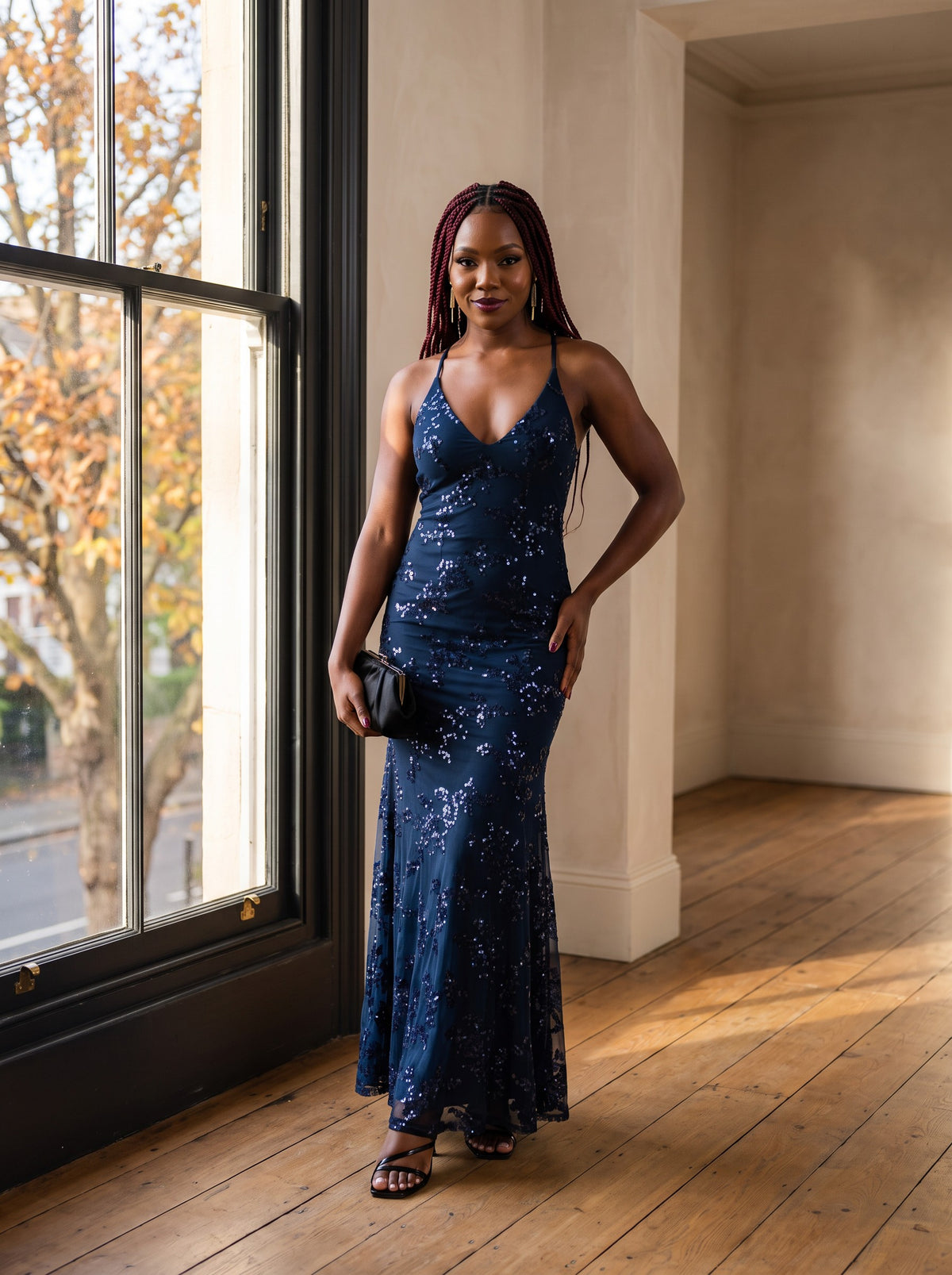 Woman in a navy sequin maxi dress by a tall window with autumn trees outside